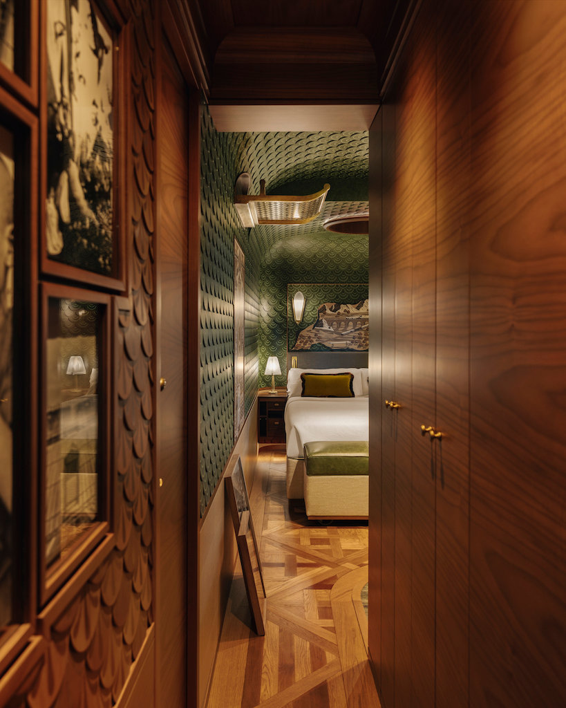 Narrow hallway leads to a cozy bedroom with green patterned walls, wood floors, and warm lighting. Framed art lines the walls. Peaceful vibe.