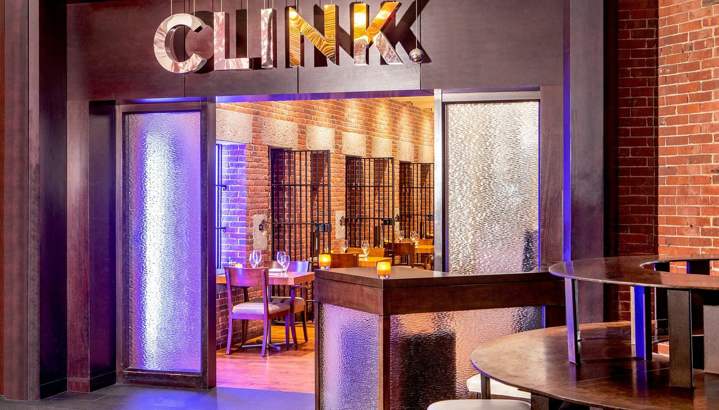 Restaurant entrance with "CLINK" sign, brick walls, and wooden tables set with candles in a cozy dining area. Warm, inviting ambiance.