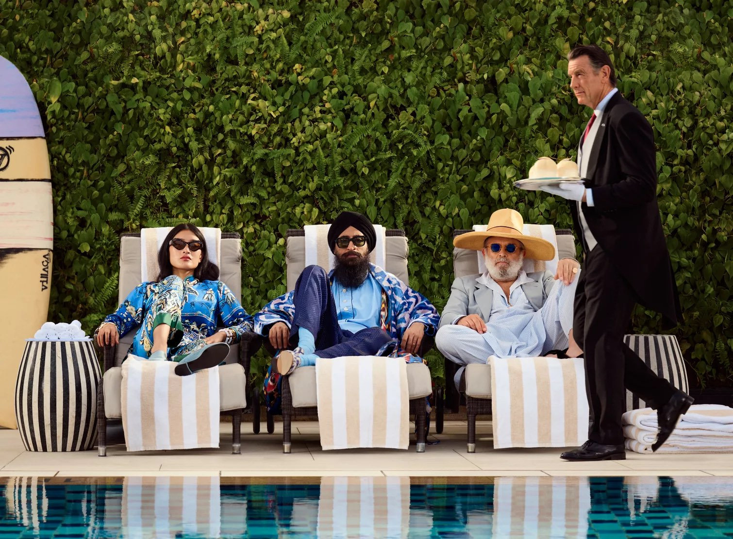 Three people in colorful attire relax on poolside loungers. A server in a suit carries a tray with drinks. Green foliage backdrop.