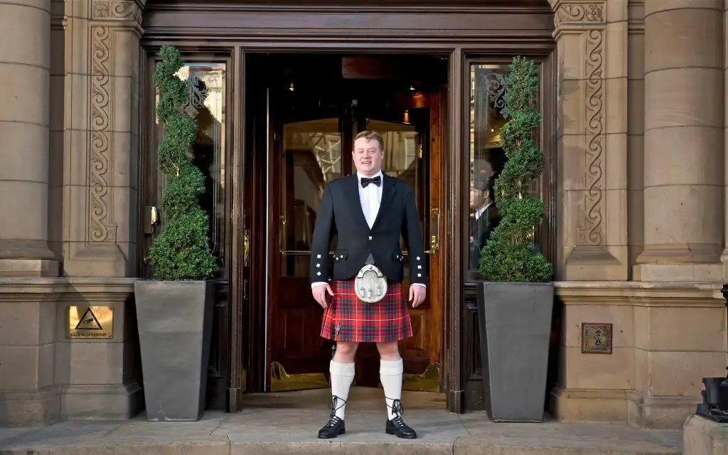 A man in a kilt and tuxedo stands smiling in a hotel entrance. Stone facade, potted plants. Elegant and welcoming atmosphere.